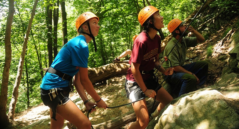 Three people wearing safety gear stand in a wooded area while looking up at an apparent rock climber. Two of them appear to be belaying the climber. 